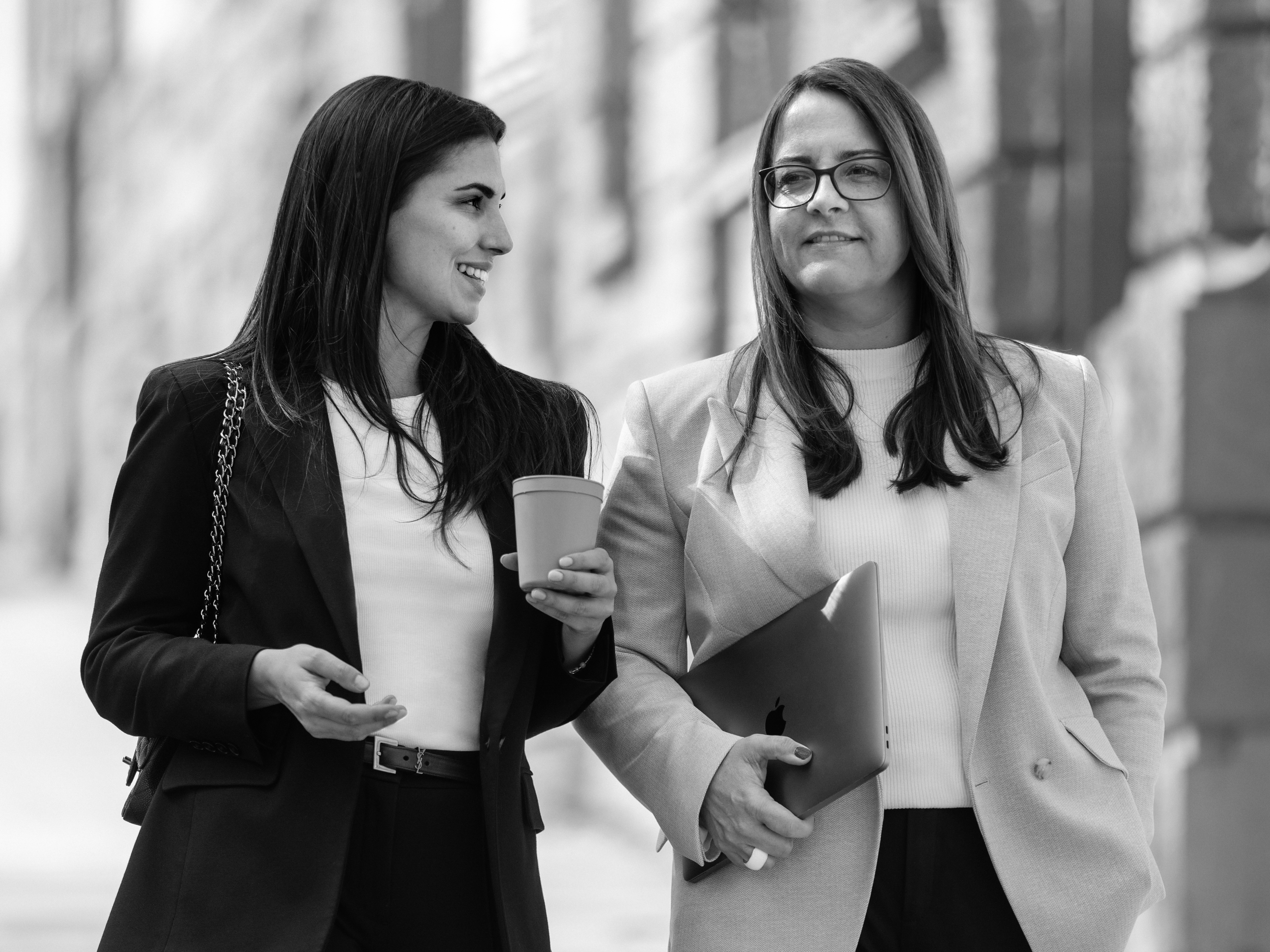 Die Rechtsanwältinnen Nadja Müller und Melanie Freiin von Neubeck unterhalten sich, während sie durch eine städtische Umgebung schlendern. Rechtsanwältin Müller hat einen Coffee to go, Rechtsanwältin von Neubeck hält ein Laptop unter ihrem Arm.  